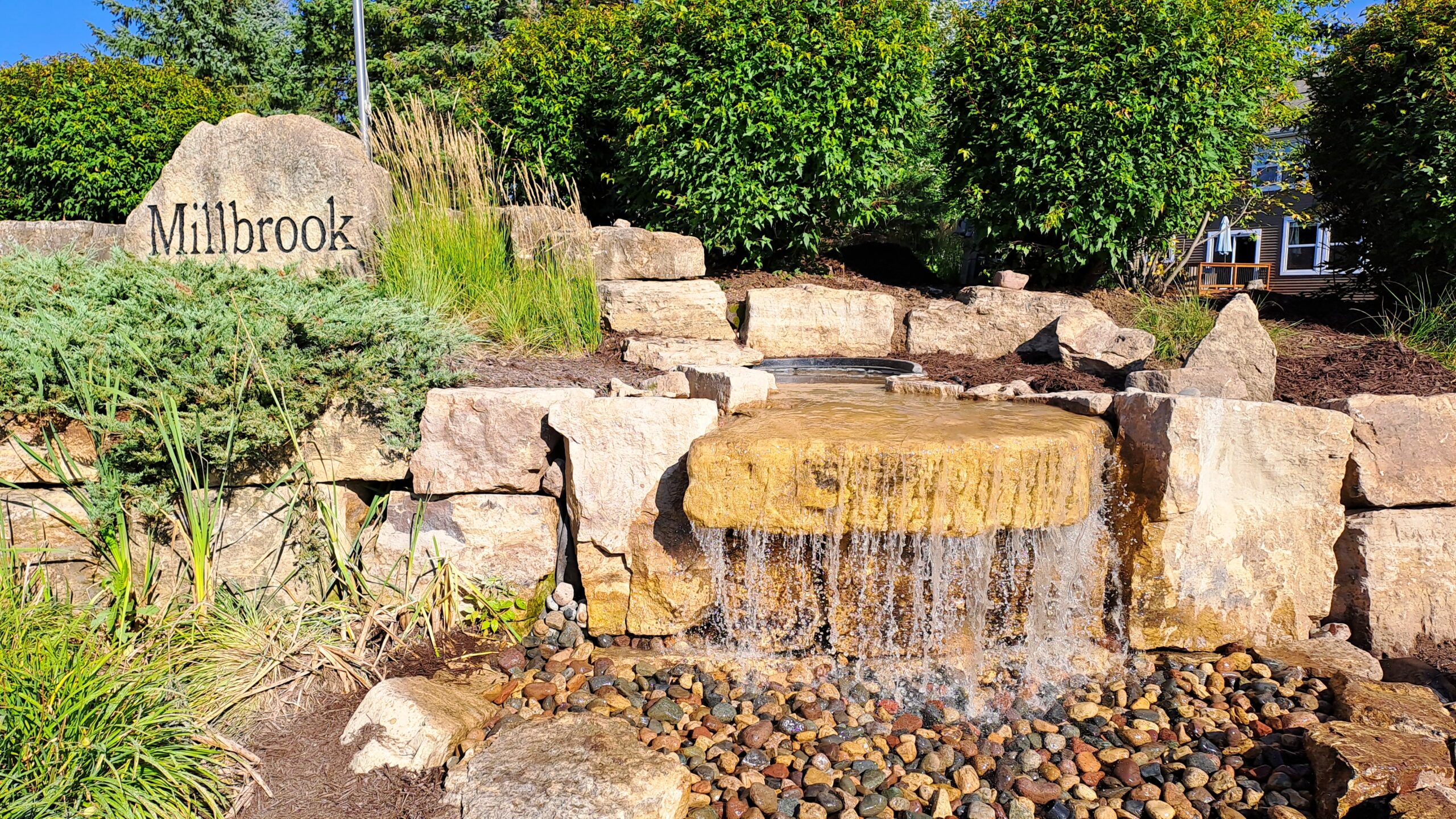 20230815_172114 Entrance pondless waterfall in Millbrook, MN with a wide limestone waterfall, community entrance sign, natural landscaping, and low-maintenance public water feature.