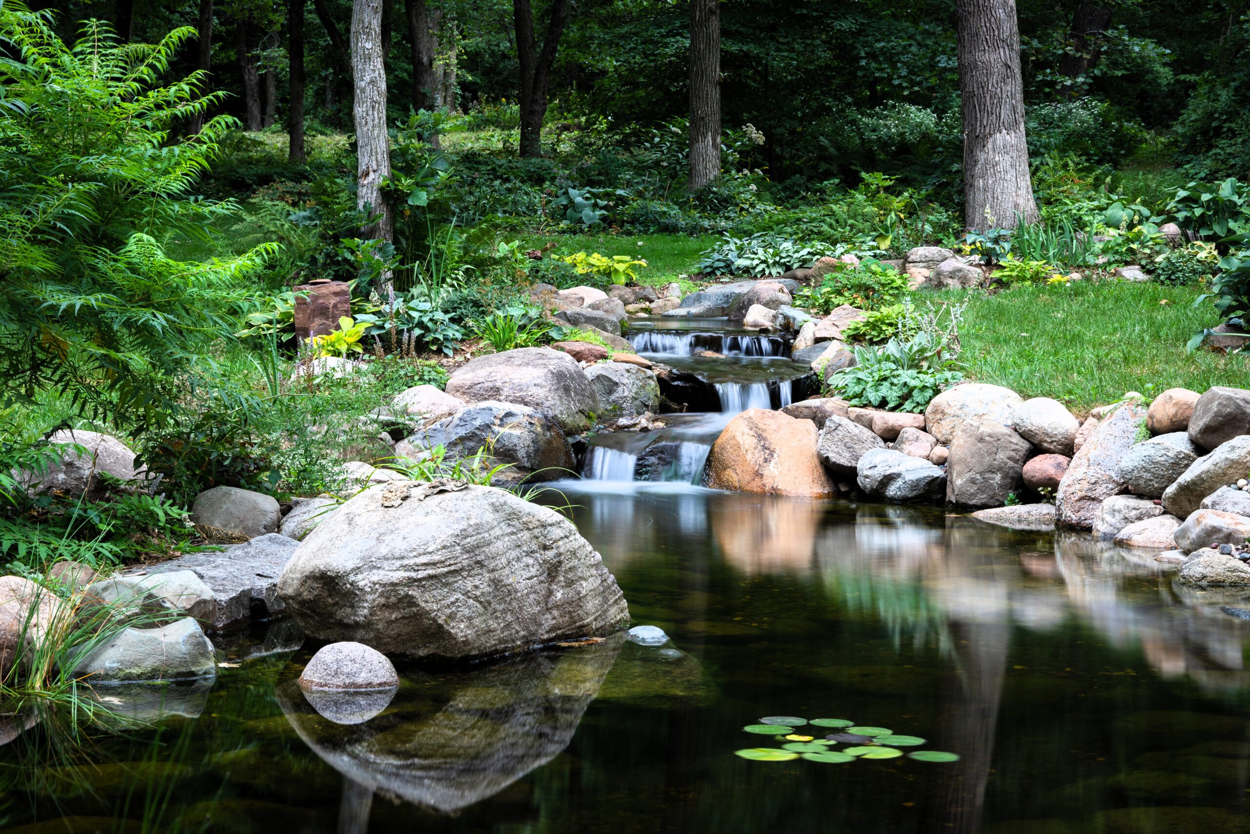 Natural backyard pond and stream in Roberts, Wisconsin with cascading stone waterfalls, clear water, natural boulders, woodland landscaping, and aquatic plants.