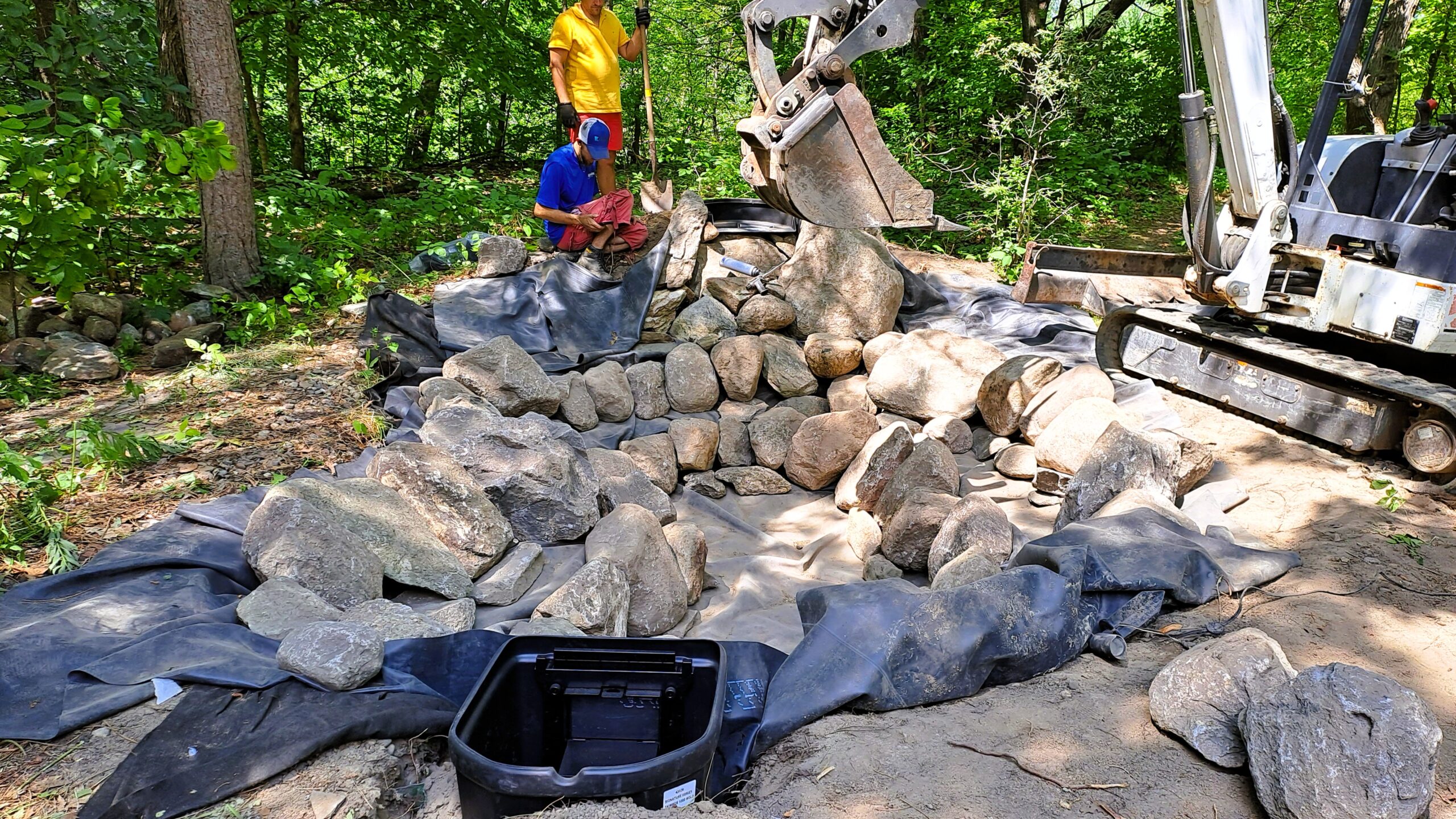 Setting waterfall boulders with excavator and shaping stream flow in backyard pond construction in Minnesota