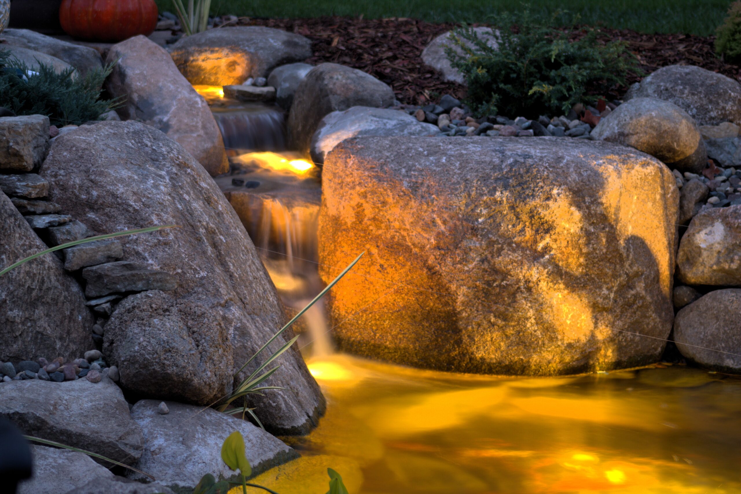Underwater LED lights illuminating a backyard pond waterfall and natural stone features at dusk in St. Michael, Minnesota