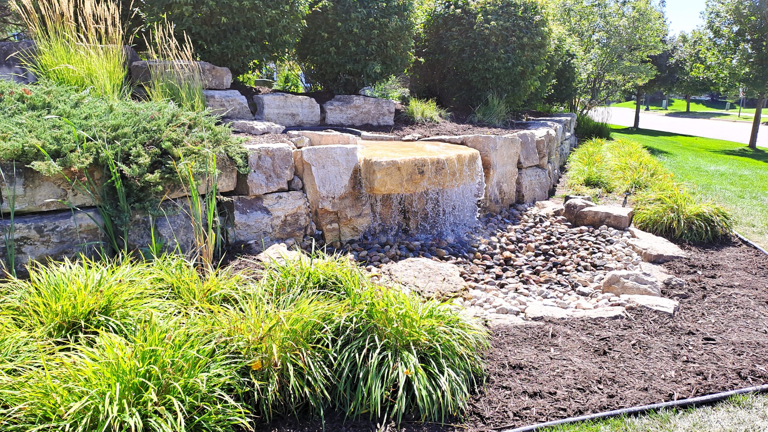 Side view of a pondless entrance waterfall in Millbrook, MN featuring natural limestone, decorative landscaping, and a low-maintenance community entrance water feature.