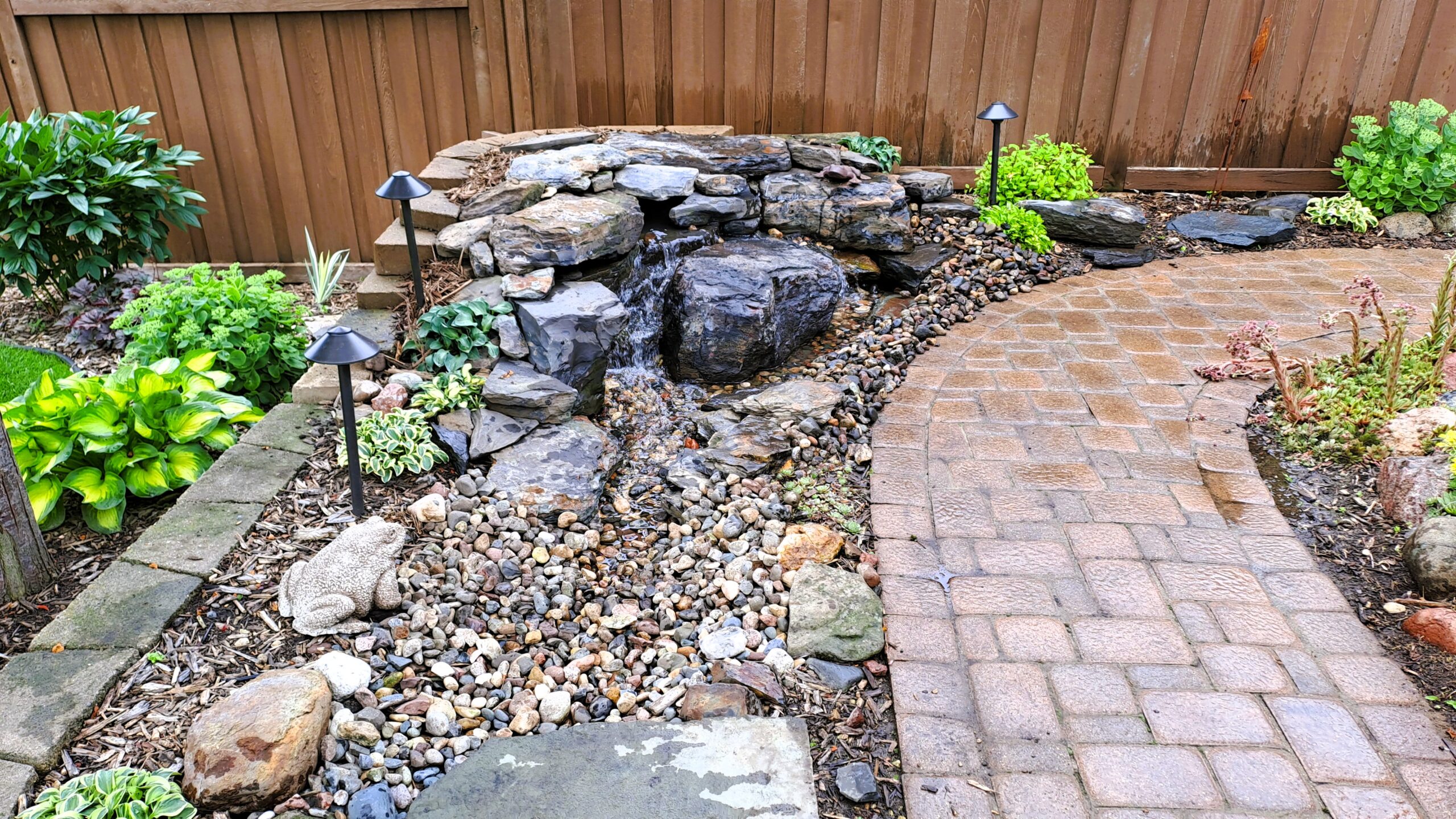 Wide-angle view of a compact pondless waterfall with Banded Jasper stone, decorative gravel stream, landscape lighting, and paver patio in Mendota Heights, MN.