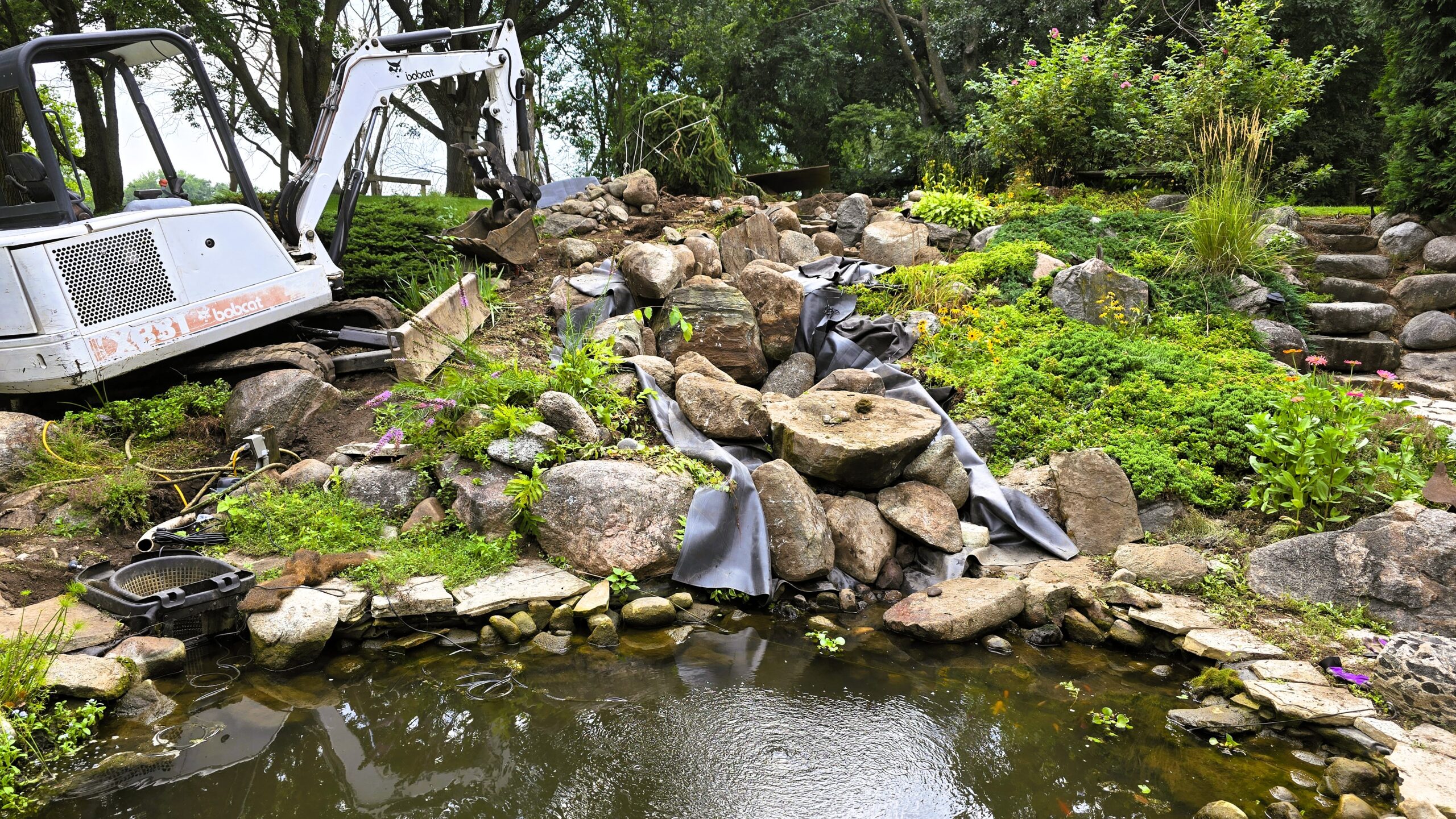 Backyard stream reconstruction in progress showing excavator, exposed liner, and natural boulders during a 35-foot stream rebuild in Elk River, MN.