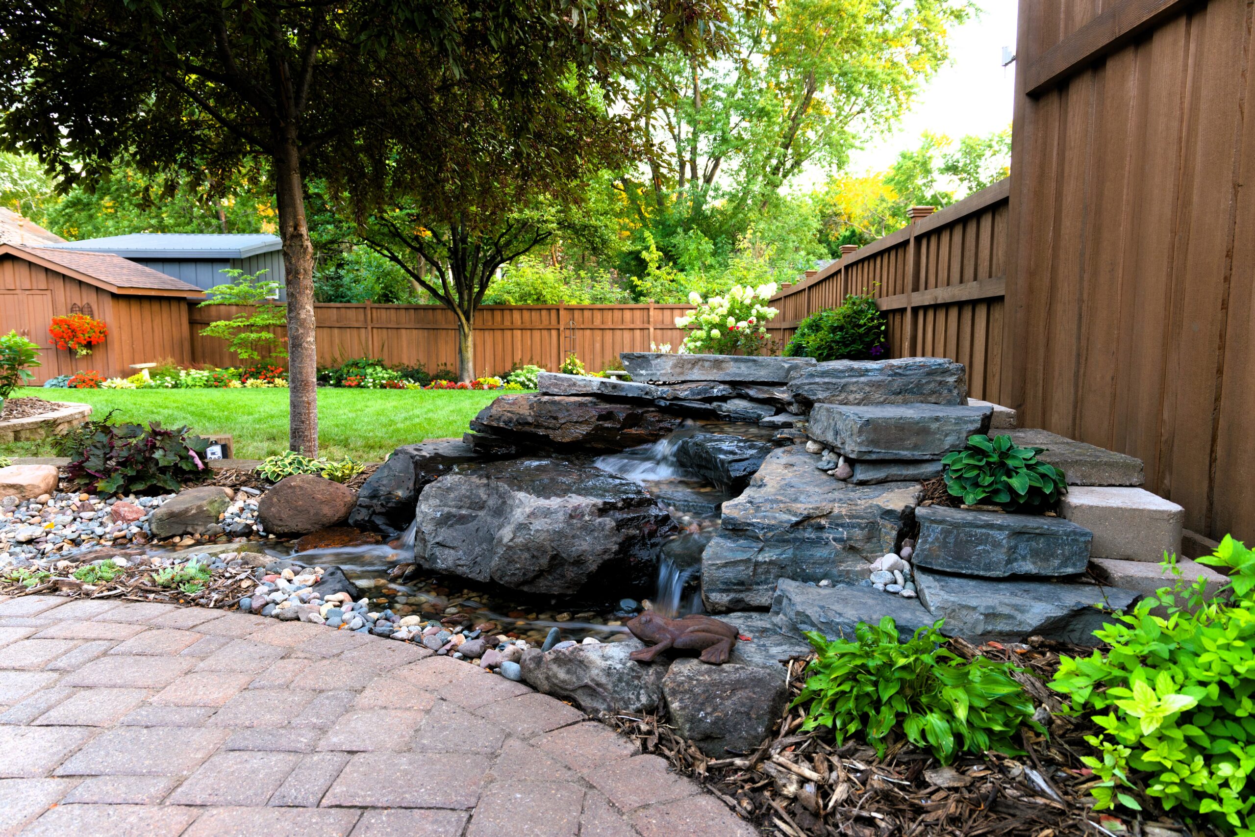 Wide-angle view of a compact pondless waterfall with Banded Jasper stone, decorative gravel stream, landscape lighting, and paver patio in Mendota Heights, MN.
