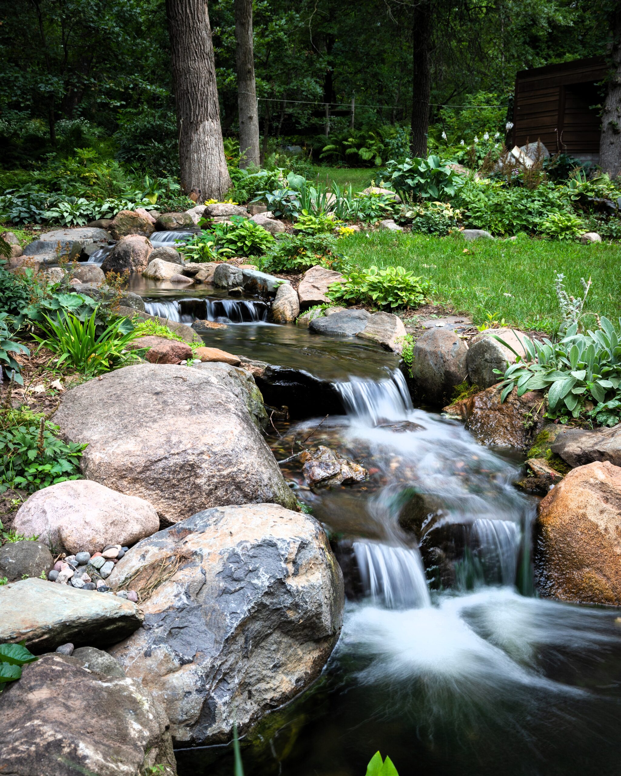 Backyard pond and stream in Roberts, Wisconsin with natural stone borders, lush landscaping, woodland setting, and clear water flowing into the pond.