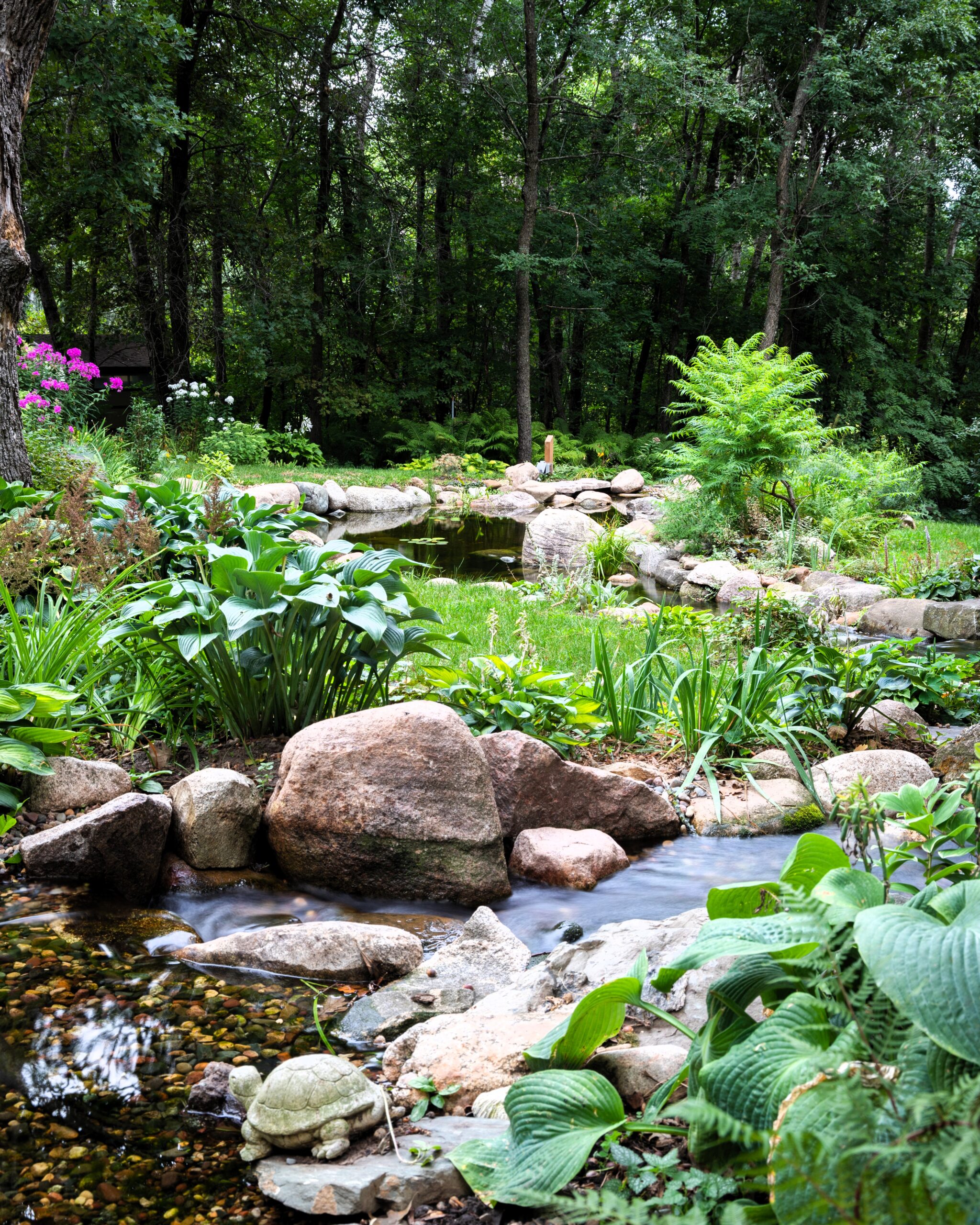 Backyard pond and stream in Roberts, Wisconsin with natural stone borders, lush landscaping, woodland setting, and clear water flowing into the pond.